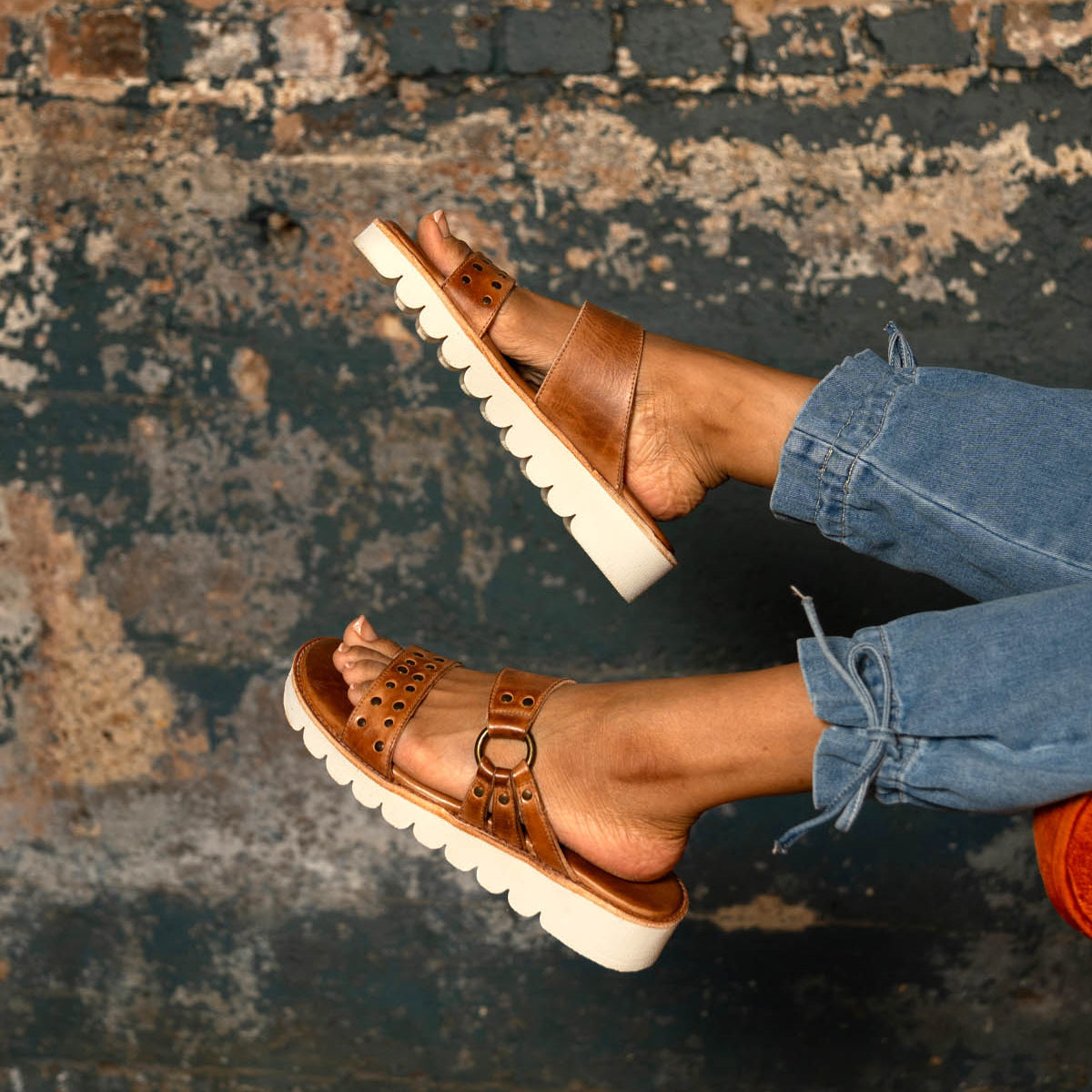 A person wearing Bed Stu's Chow brown leather slide sandals with a chunky rubber sole and blue jeans stands in front of a textured, weathered wall.