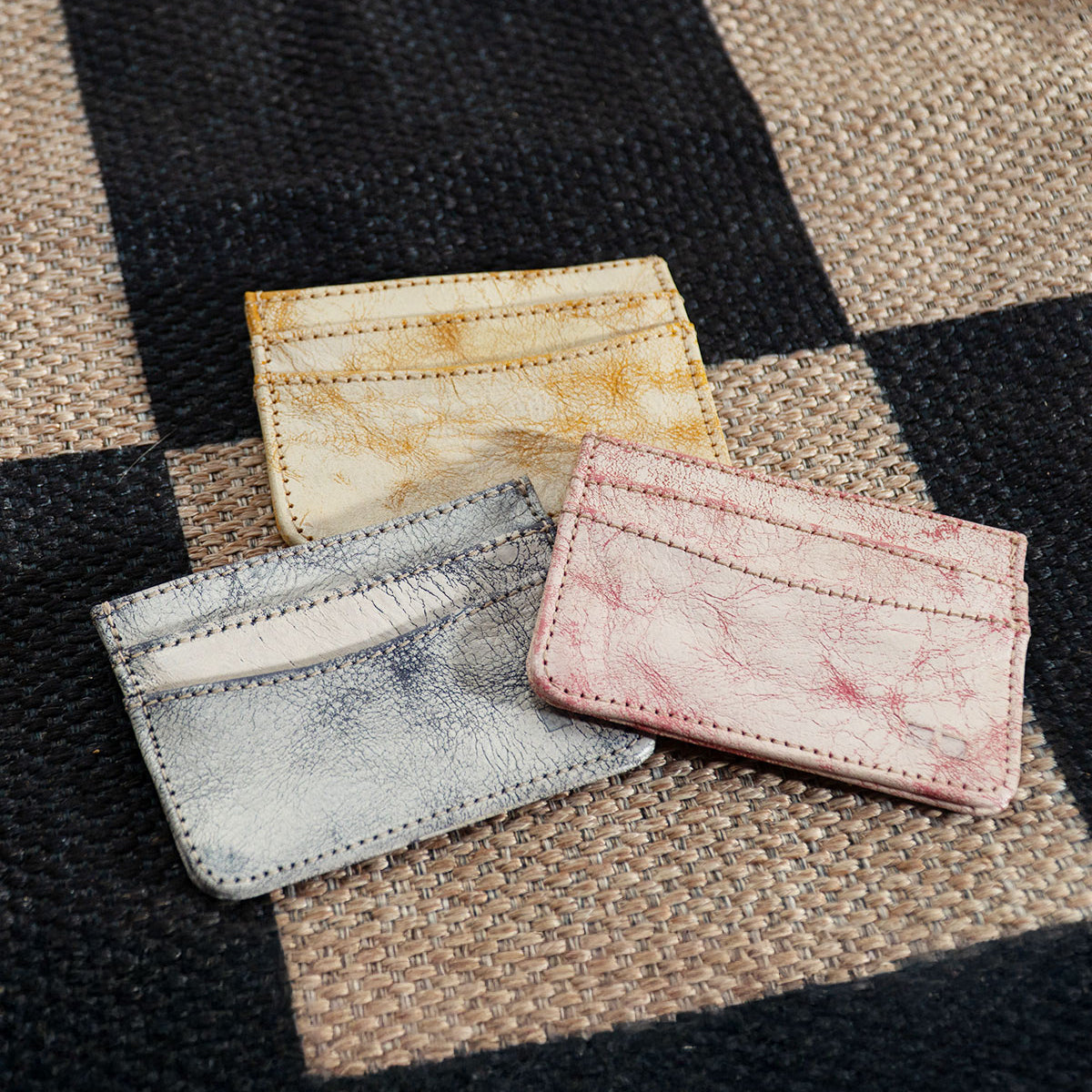 Three Chuck leather card holders by Bed Stu in yellow, pink, and gray rest elegantly on a woven checkered fabric background—ideal for organizing your credit cards or business cards with style.
