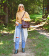 A blonde woman wearing a yellow sweater and jeans standing in a wooded area with a Bed Stu Cirdan leather backpack featuring adjustable straps and a zip-top closure.