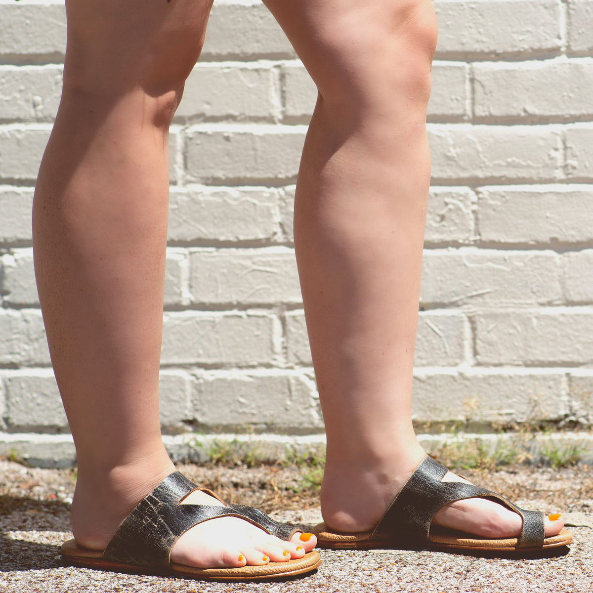 Person wearing black Civilized sandals by Bed Stu with a cushioned sole and standing on pavement with legs crossed in front of a white brick wall, exuding a laid-back style.