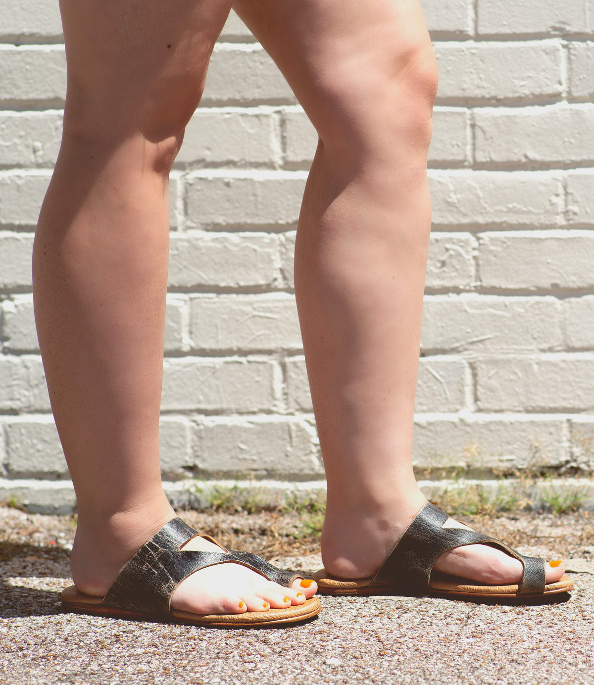 Person wearing black Civilized sandals by Bed Stu with a cushioned sole and standing on pavement with legs crossed in front of a white brick wall, exuding a laid-back style.