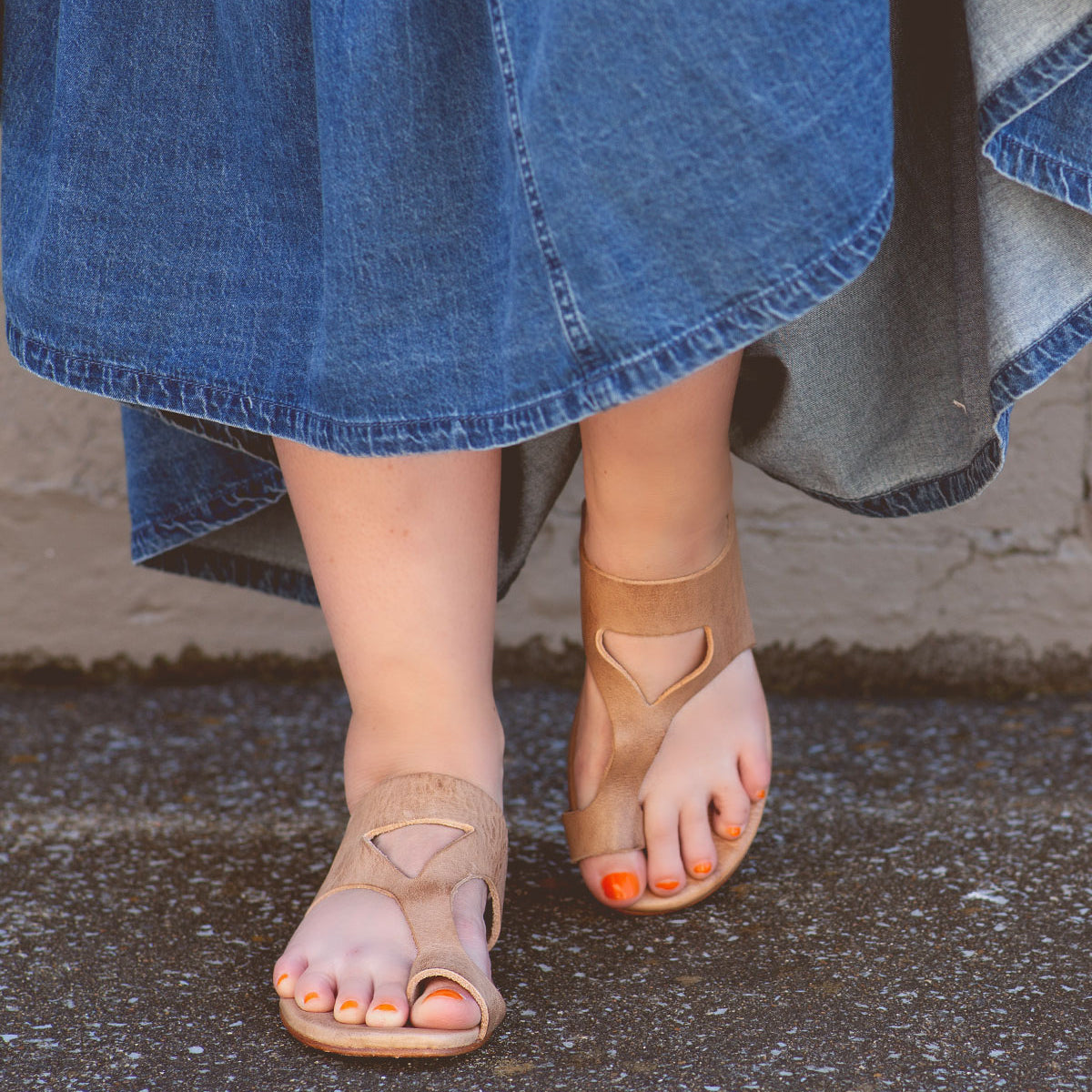 Person wearing a long denim skirt and tan Bed Stu sandals with a unique design called Civilized, featuring cushioned soles for added comfort, standing on an asphalt surface. The person's toenails are painted orange, adding to the laid-back style.