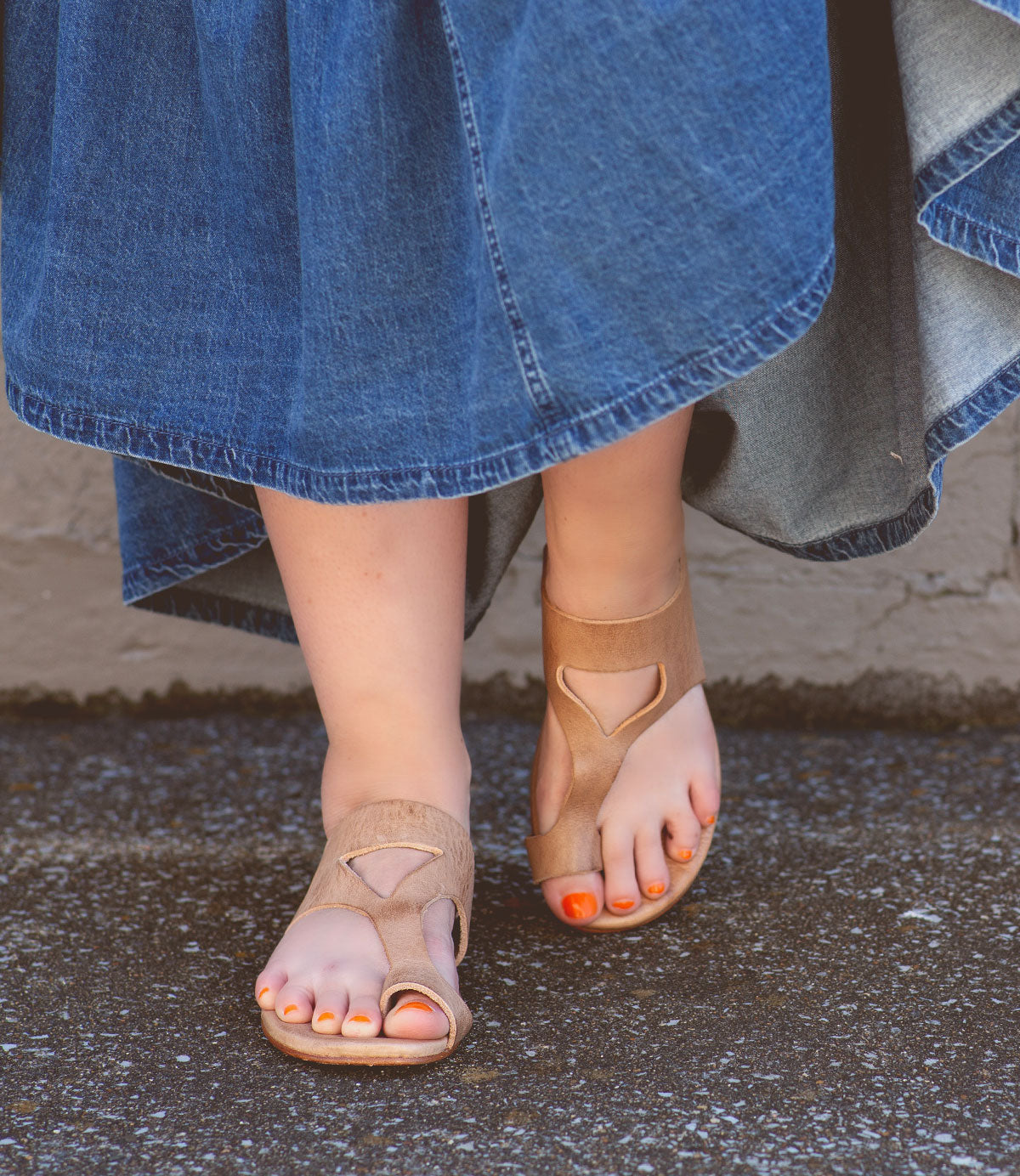 Person wearing a long denim skirt and tan Bed Stu sandals with a unique design called Civilized, featuring cushioned soles for added comfort, standing on an asphalt surface. The person's toenails are painted orange, adding to the laid-back style.