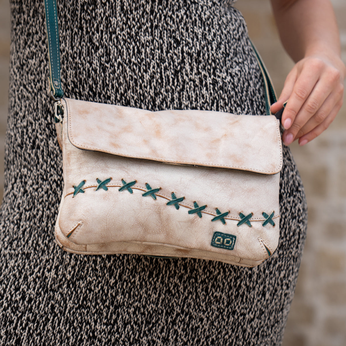 A person holds the Bed Stu Cleo, a beige crossbody bag with green stitching and zipper top, while wearing a grey textured dress outdoors.