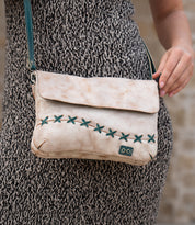 A person holds the Bed Stu Cleo, a beige crossbody bag with green stitching and zipper top, while wearing a grey textured dress outdoors.