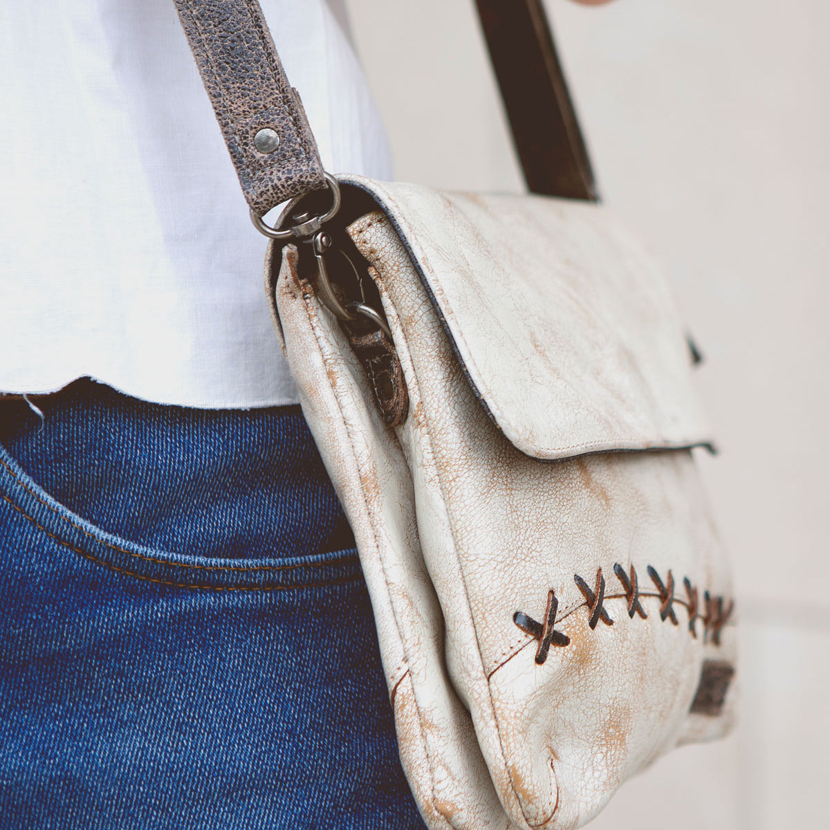 A person wearing blue jeans and a white shirt is shown from the waist down, carrying a small, distressed beige leather Bed Stu Cleo crossbody bag with dark brown straps, stitching details, and a zipper top closure.