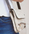 A person wearing blue jeans and a white shirt is shown from the waist down, carrying a small, distressed beige leather Bed Stu Cleo crossbody bag with dark brown straps, stitching details, and a zipper top closure.
