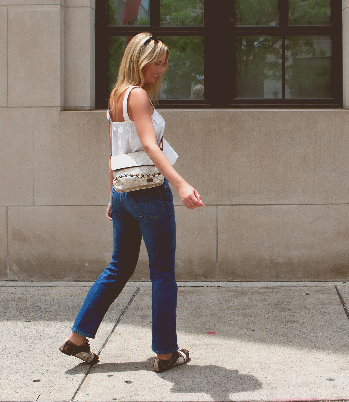 A woman with blonde hair walks on a sidewalk in front of a building, sporting a white top, blue jeans, brown sandals, and a versatile Bed Stu Cleo crossbody bag with a zipper top closure. She is looking down and slightly smiling.