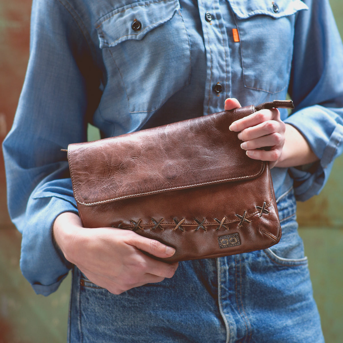 A woman holding a Bed Stu Cleo brown leather clutch with an adjustable strap.