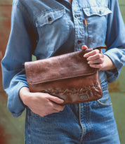 A woman holding a Bed Stu Cleo brown leather clutch with an adjustable strap.