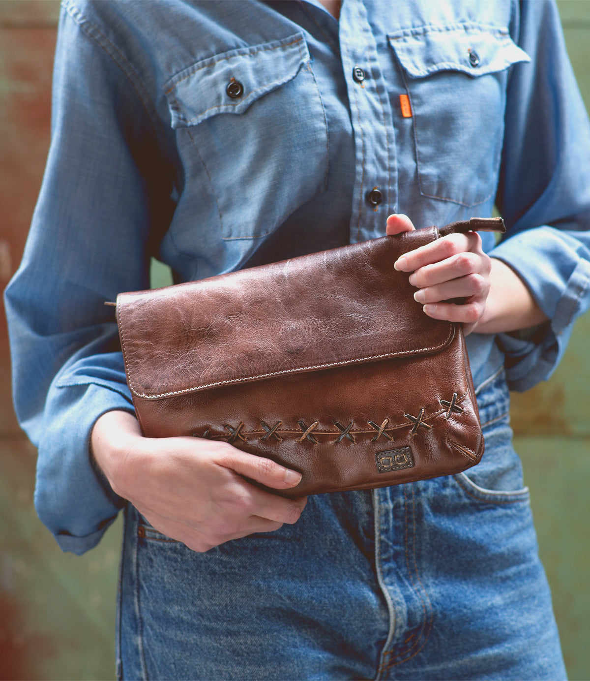 A woman holding a Bed Stu Cleo brown leather clutch with an adjustable strap.