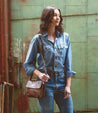 A woman wearing jeans and a denim shirt standing in front of a building, clutching a Bed Stu Cleo adjustable crossbody bag.