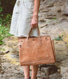 A woman is holding a multifunctional Bed Stu tan leather bag perfect for commuters with laptops.