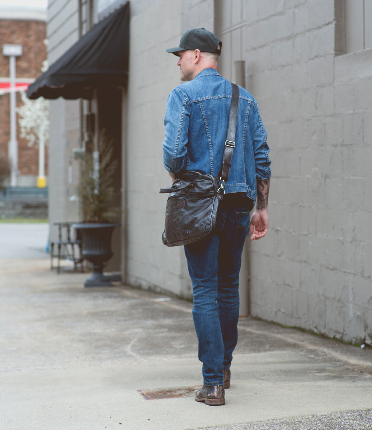 A man wearing a denim jacket and jeans walking down the sidewalk with a Bed Stu conclusion vegetable-tanned leather laptop bag.