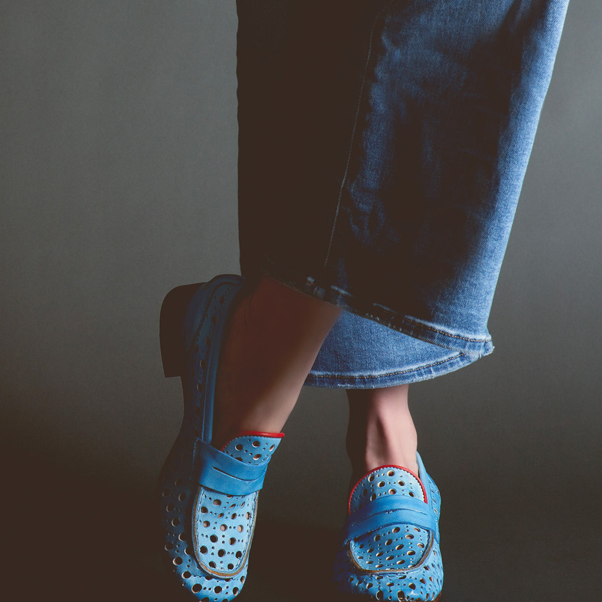 A person wearing Bed Stu Cornflower penny loafers with a circular cutout pattern and denim jeans against a dark background.