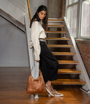A woman wearing a white sweater, black pants, and metallic leather sneakers leans against a staircase railing while holding a large brown leather Bed|Stü Cosmo hobo bag.