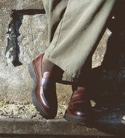 A person wearing Bed Stu's Costruito loafers with a chunky Livetech outsole and olive green pants stands against a stone wall.