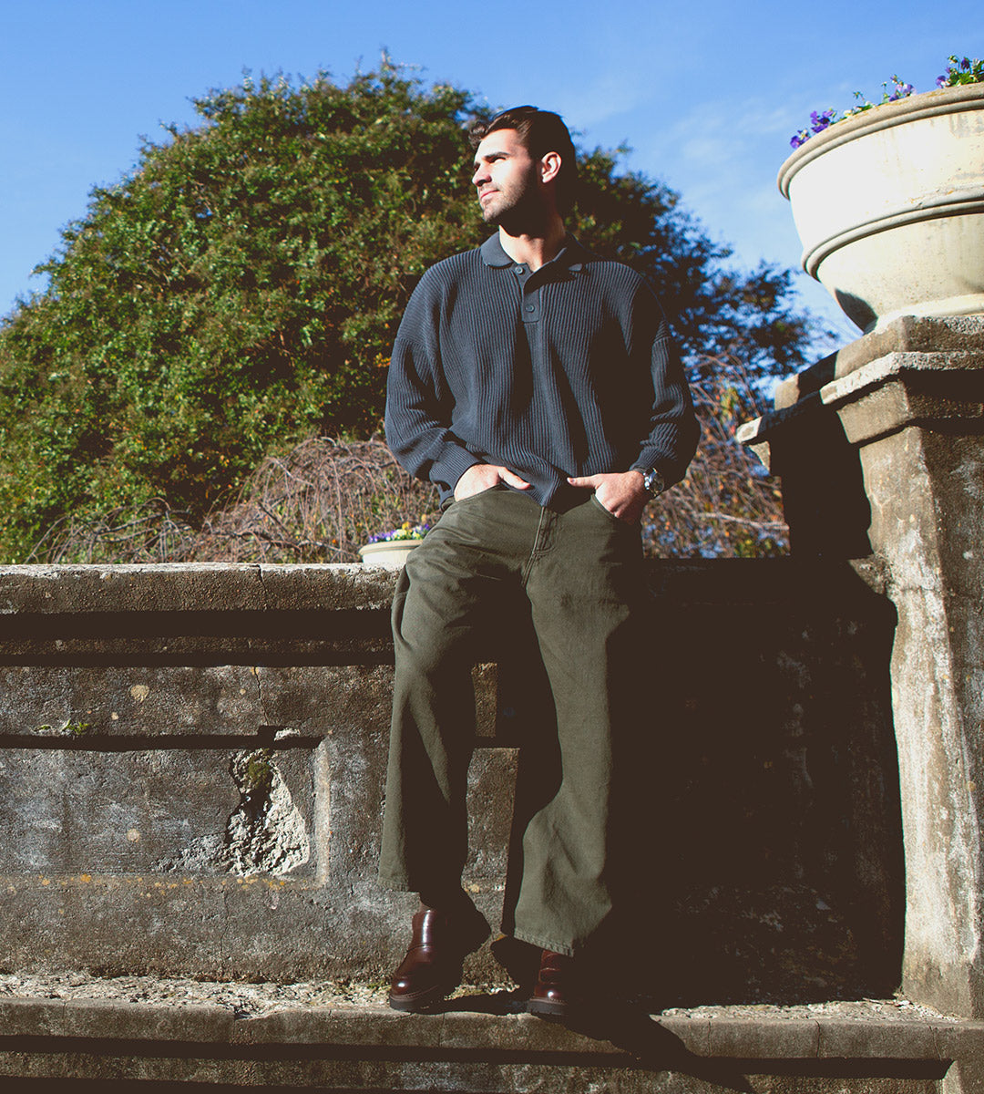 A man stands against an old stone wall, dressed in a dark sweater and green pants beneath a clear blue sky. His Costruito penny loafers by Bed Stu, crafted in Italy with a chunky Livetech outsole, elegantly enhance his relaxed look.