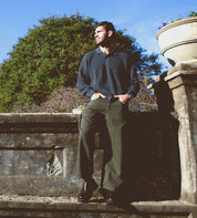 A man stands against an old stone wall, dressed in a dark sweater and green pants beneath a clear blue sky. His Costruito penny loafers by Bed Stu, crafted in Italy with a chunky Livetech outsole, elegantly enhance his relaxed look.