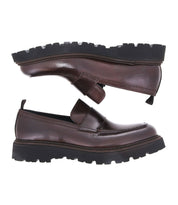 The Costruito penny loafers by Bed Stu, crafted in Italy from brown leather and boasting thick black rubber soles, are displayed from a side view. These shoes combine artisanal charm with a sophisticated design, ideal for those who value fine craftsmanship.