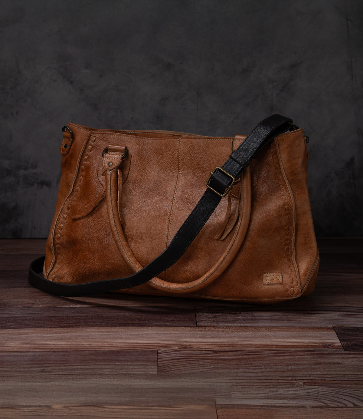 A Bed Stu Crossbody Strap in brown leather with dual top handles and a black adjustable strap sits on a wooden floor against a dark textured background.