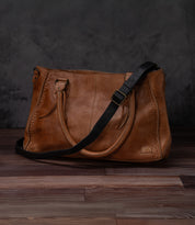 A Bed Stu Crossbody Strap in brown leather with dual top handles and a black adjustable strap sits on a wooden floor against a dark textured background.