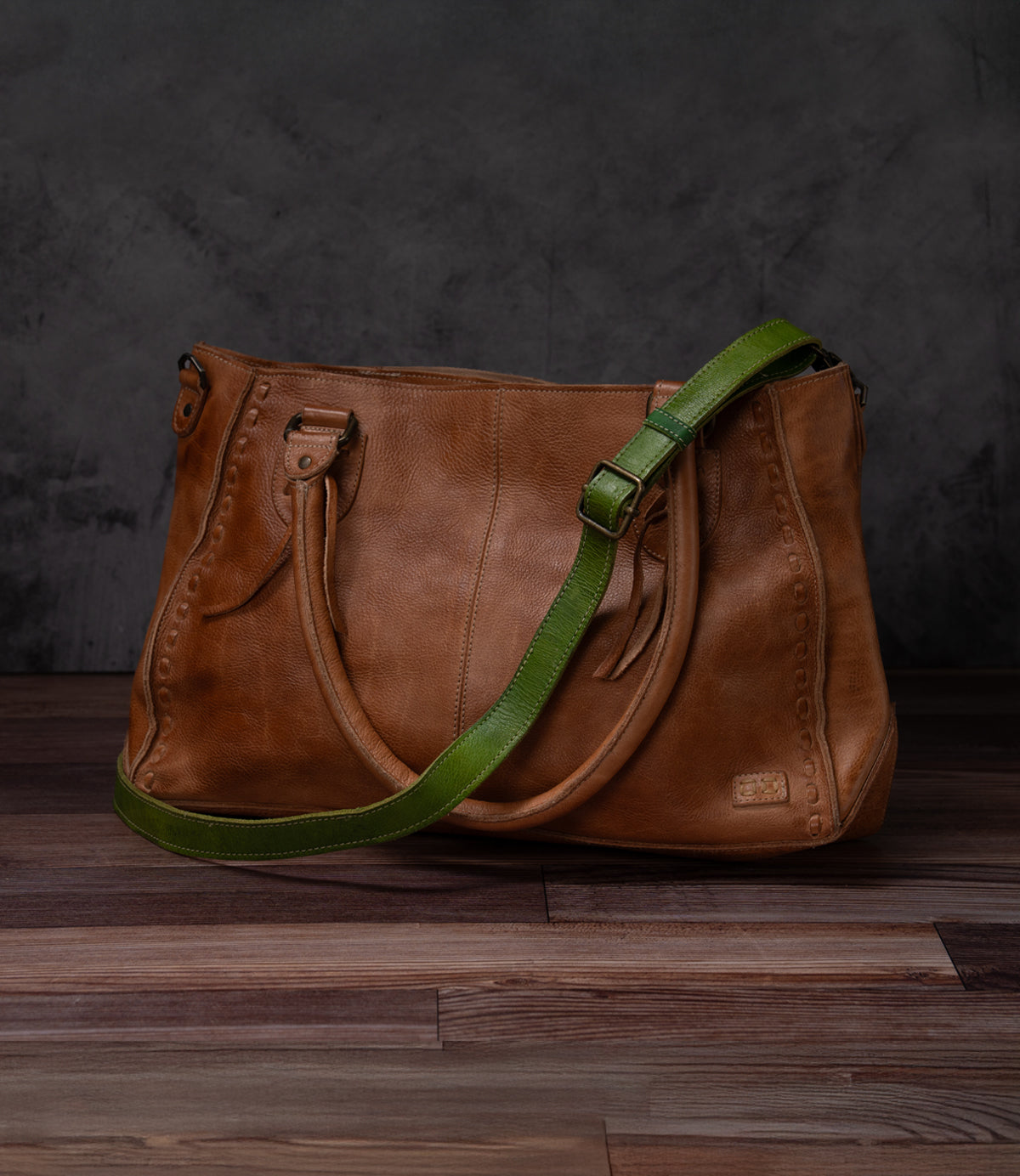 A Bed Stu Crossbody Strap with double handles and an adjustable leather strap rests on a wooden floor against a dark textured background, ideal for wearing as a crossbody.