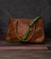 A Bed Stu Crossbody Strap with double handles and an adjustable leather strap rests on a wooden floor against a dark textured background, ideal for wearing as a crossbody.