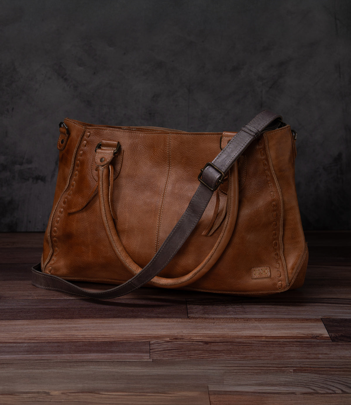 A Bed Stu Crossbody Strap in brown leather with double handles and an adjustable strap is displayed on a wooden floor against a dark background.