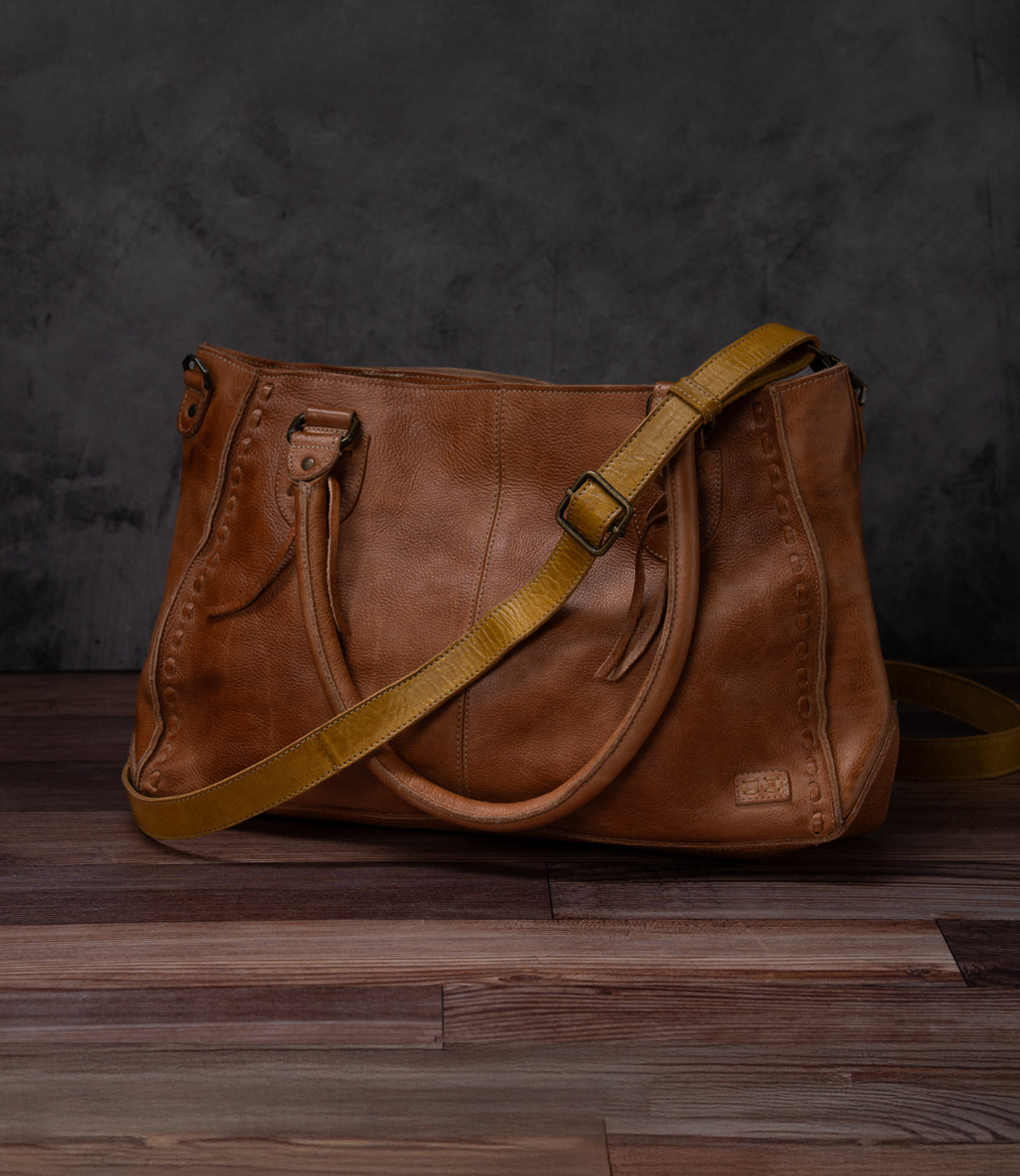 A Bed Stu Crossbody Strap in brown leather features double handles and an adjustable strap, displayed on a wooden surface with a dark background.