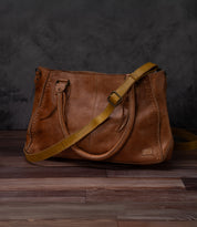 A Bed Stu Crossbody Strap in brown leather features double handles and an adjustable strap, displayed on a wooden surface with a dark background.