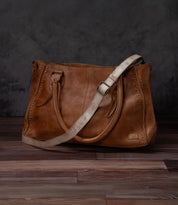 A Bed Stu Crossbody Strap bag with dual handles and an adjustable leather strap rests on a wooden floor before a dark textured backdrop.
