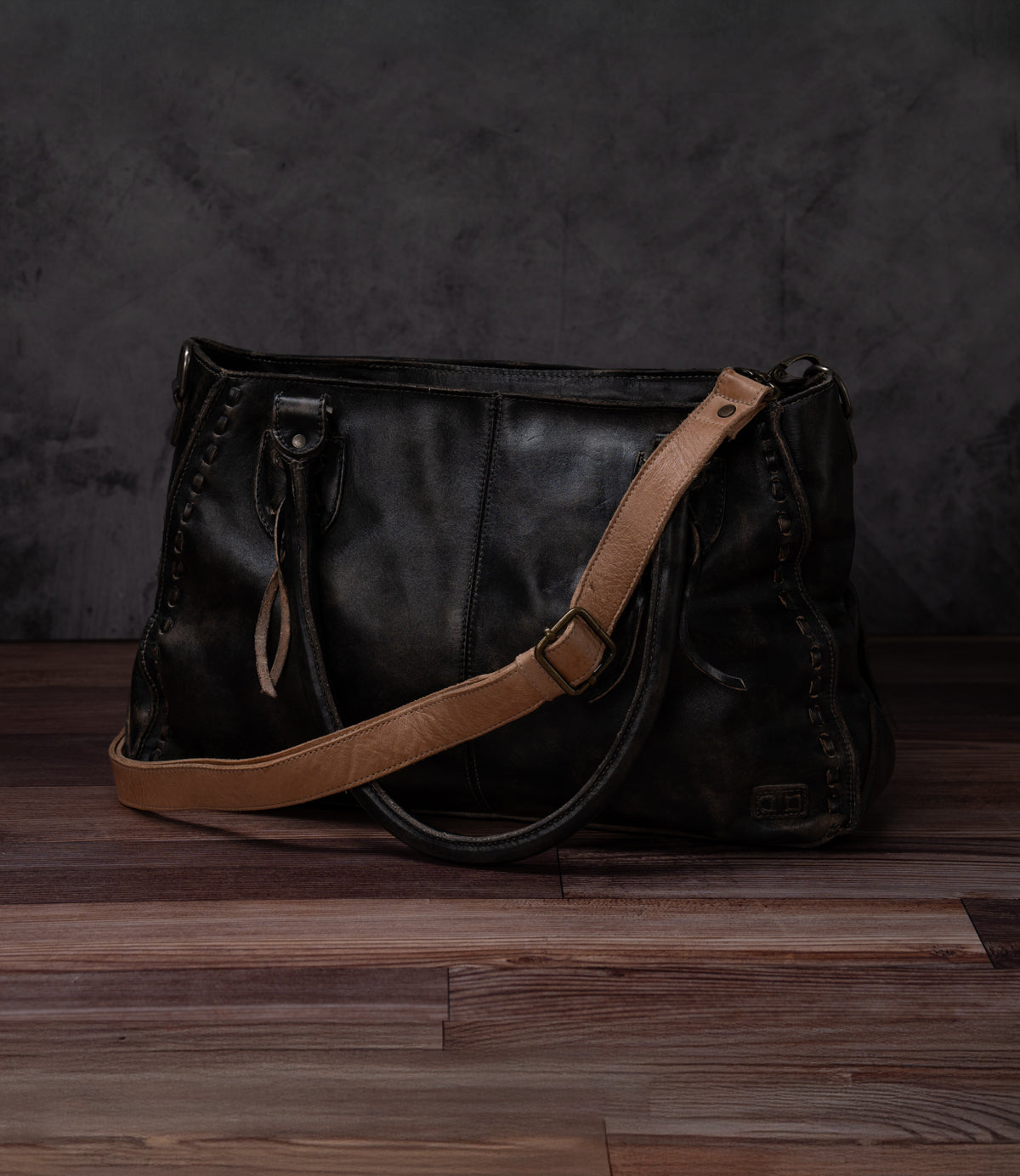 A Bed Stu Crossbody Strap in black leather with a tan adjustable strap rests on a wooden floor against a dark textured backdrop.
