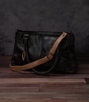 A Bed Stu Crossbody Strap in black leather with a tan adjustable strap rests on a wooden floor against a dark textured backdrop.