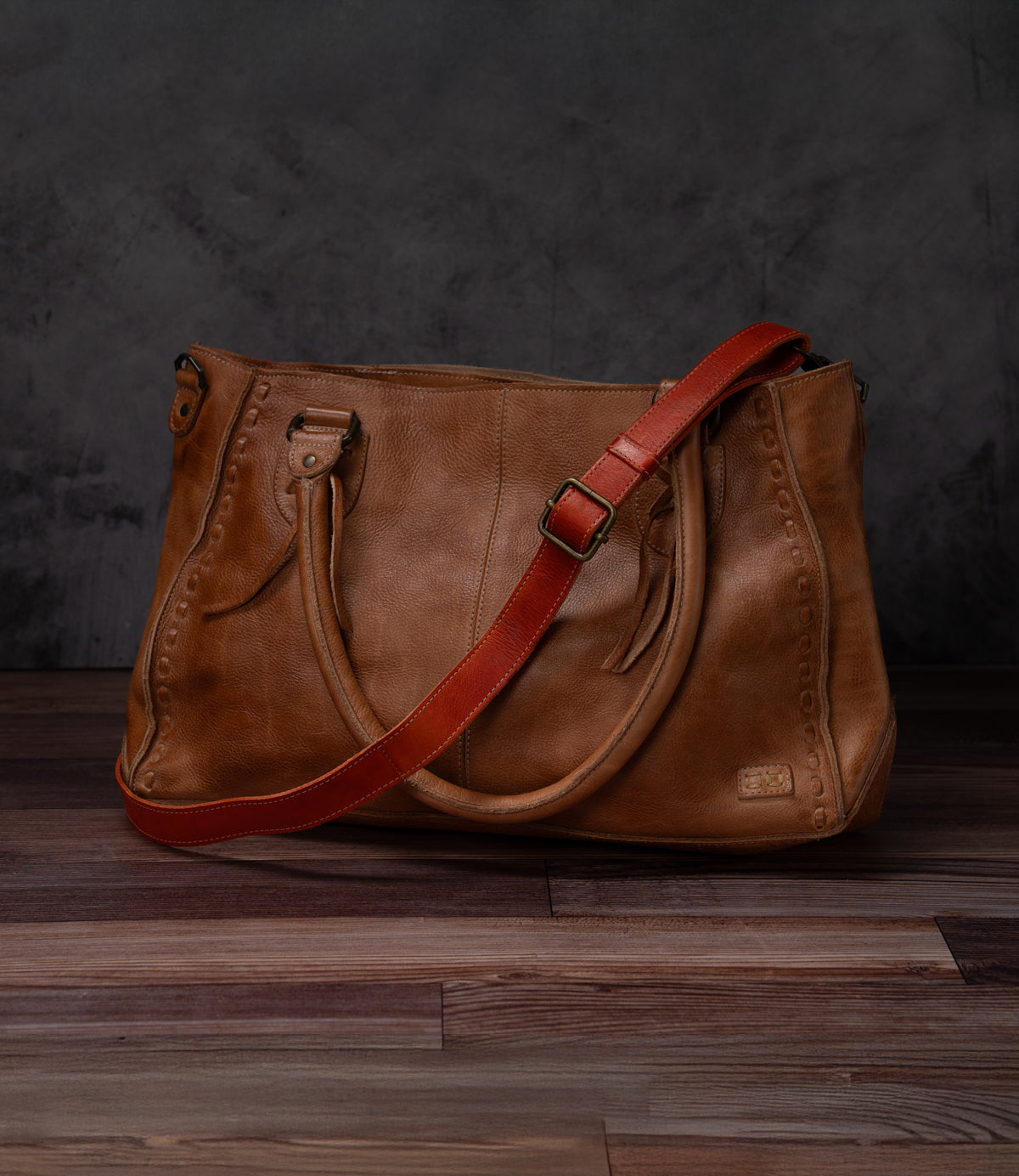 A Bed Stu Crossbody Strap in brown leather, featuring two handles and a red adjustable strap for crossbody wear, rests on a wooden floor with a dark, textured backdrop.