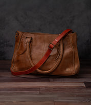 A Bed Stu Crossbody Strap in brown leather, featuring two handles and a red adjustable strap for crossbody wear, rests on a wooden floor with a dark, textured backdrop.