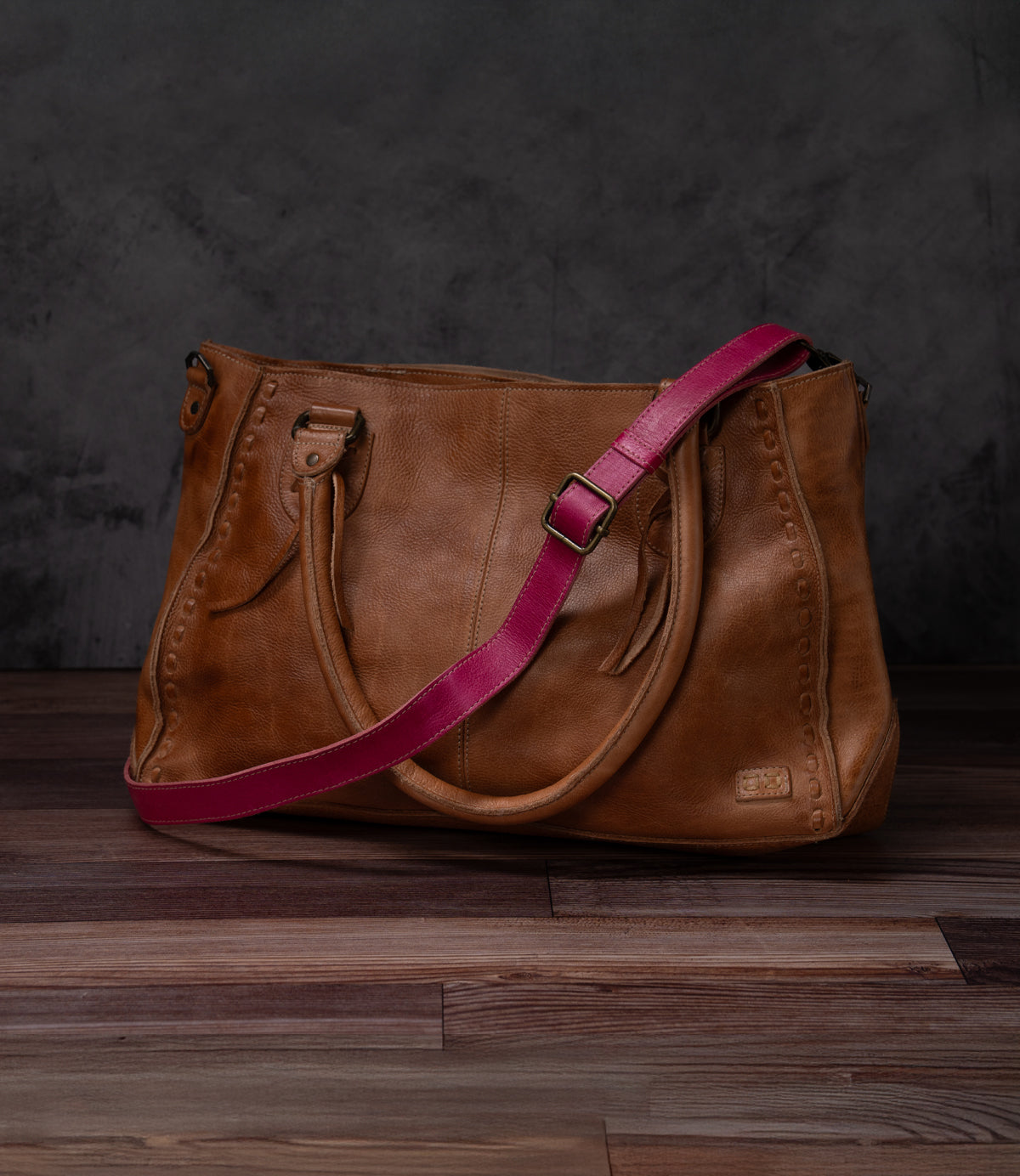 A Bed Stu Crossbody Strap in brown leather with two handles and a pink adjustable strap, designed for crossbody use, shown on a wooden floor against a dark textured background.