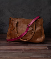 A Bed Stu Crossbody Strap in brown leather with two handles and a pink adjustable strap, designed for crossbody use, shown on a wooden floor against a dark textured background.