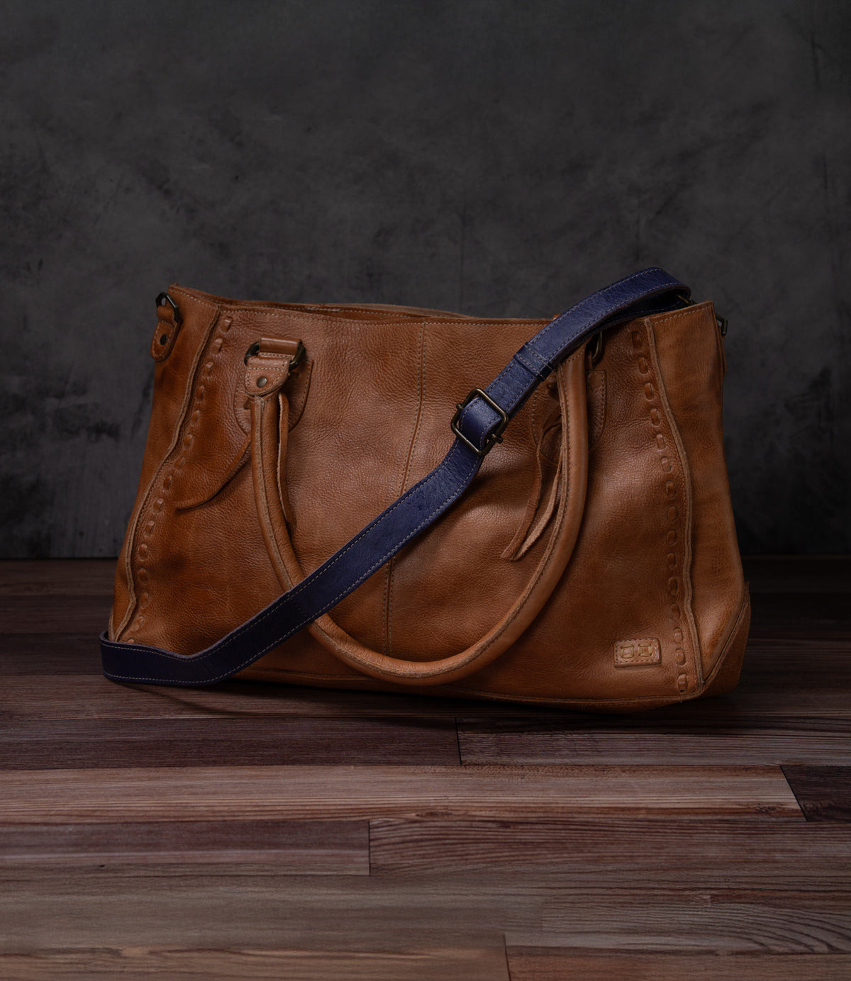 A Bed Stu Crossbody Strap with dual handles and a dark blue adjustable leather strap rests on a wooden floor against a dark, textured background.
