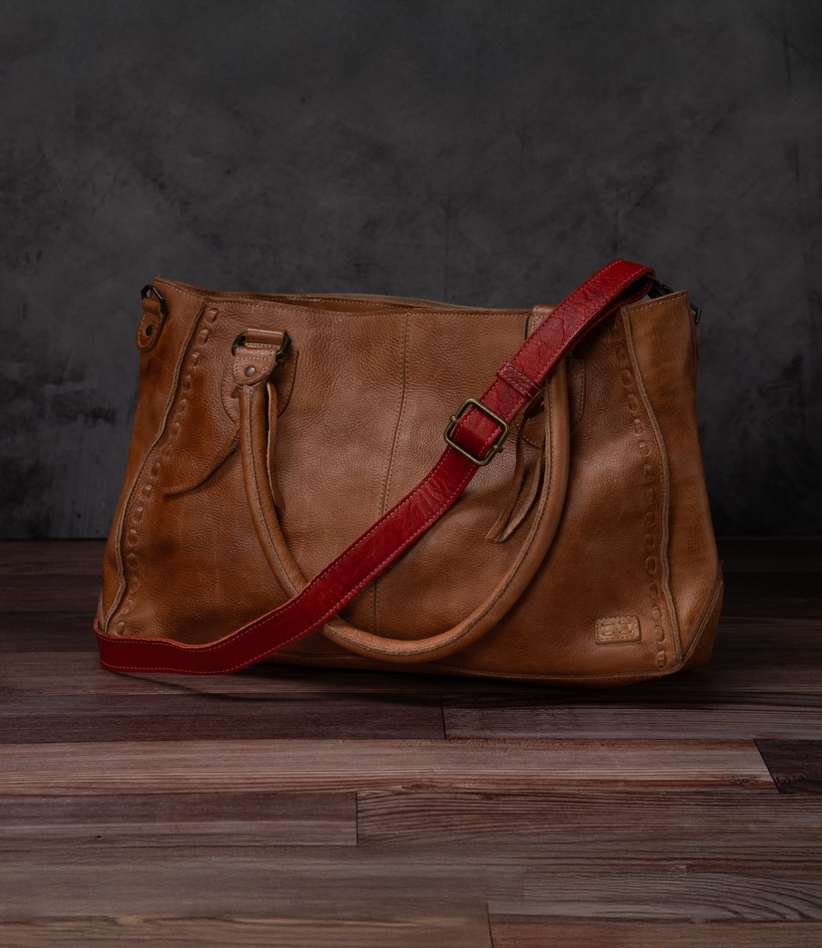 A Bed Stu Crossbody Strap bag in brown with two handles and an adjustable leather strap, ideal for crossbody wear, is shown on a wooden floor against a dark background.