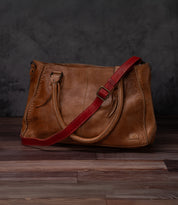 A Bed Stu Crossbody Strap bag in brown with two handles and an adjustable leather strap, ideal for crossbody wear, is shown on a wooden floor against a dark background.