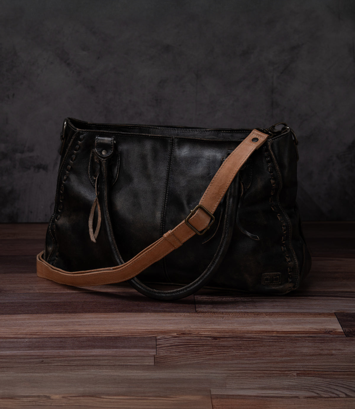 A Bed Stu Crossbody Strap in black features a tan adjustable leather strap and is displayed on a wooden floor against a dark, textured background.