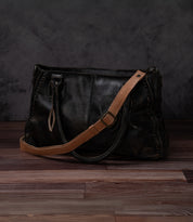 A Bed Stu Crossbody Strap in black features a tan adjustable leather strap and is displayed on a wooden floor against a dark, textured background.