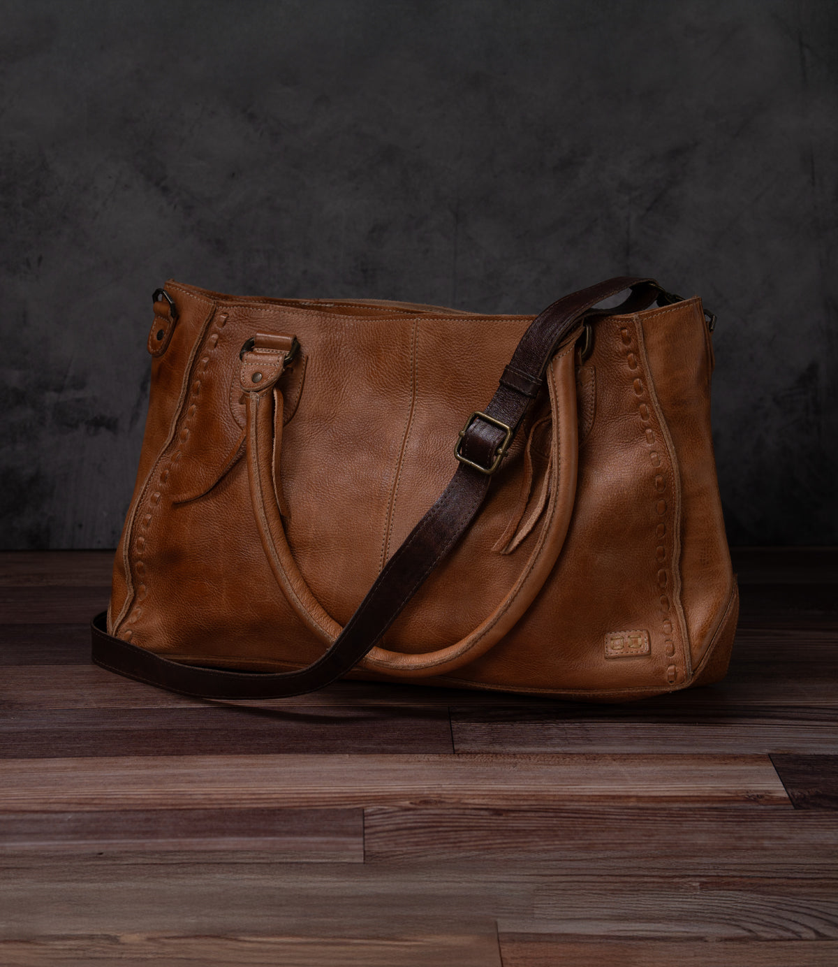 A Bed Stu Crossbody Strap bag in brown leather with dual top handles and a dark brown adjustable strap, shown on a wooden floor against a dark textured background.