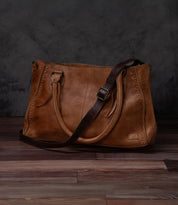 A Bed Stu Crossbody Strap bag in brown leather with dual top handles and a dark brown adjustable strap, shown on a wooden floor against a dark textured background.