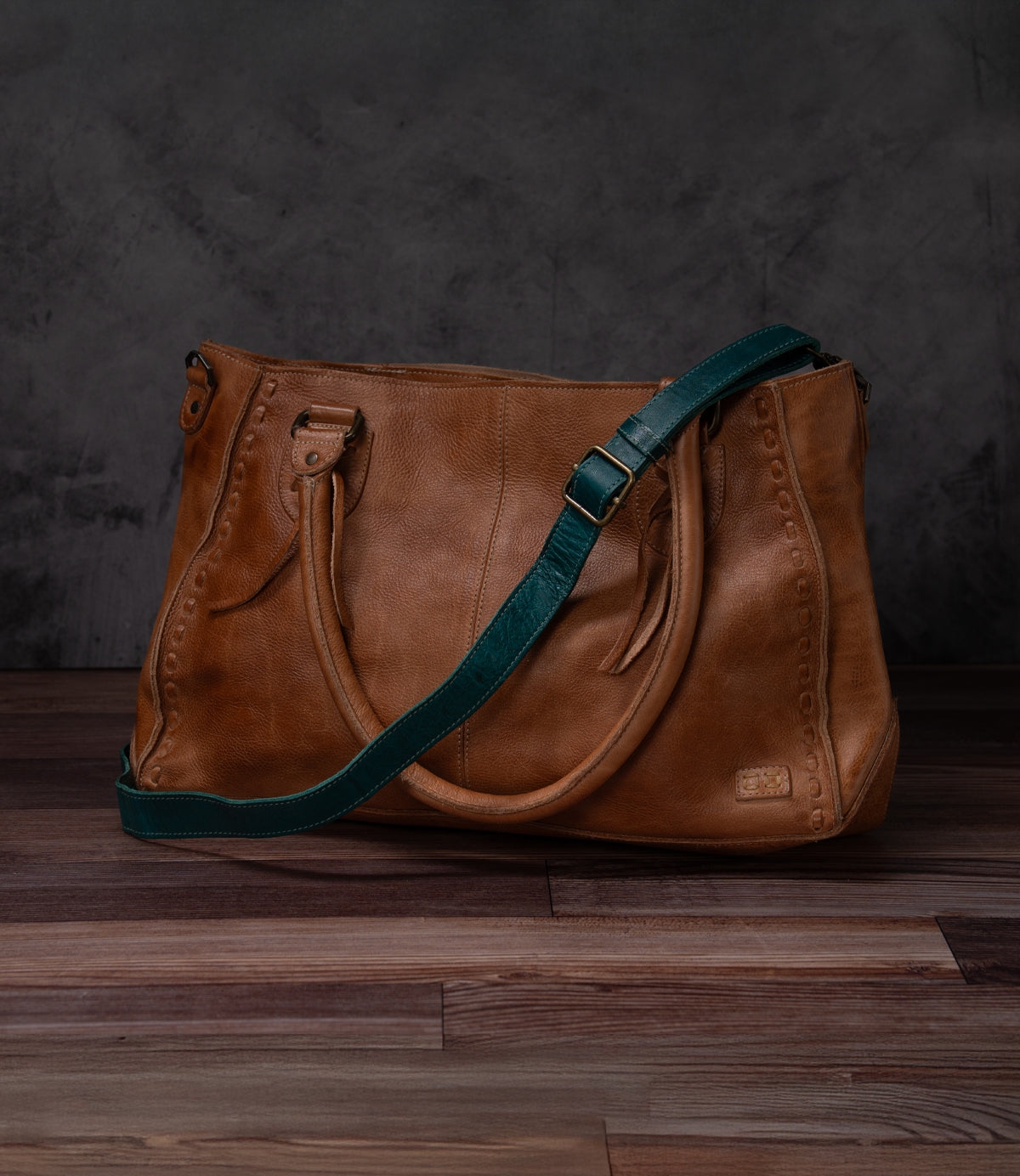 Brown leather Crossbody Strap by Bed Stu, featuring decorative stitching, double handles, and an adjustable strap for crossbody wear, shown on a wooden floor with a dark textured backdrop—showcasing Bed Stu's signature style.