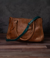 Brown leather Crossbody Strap by Bed Stu, featuring decorative stitching, double handles, and an adjustable strap for crossbody wear, shown on a wooden floor with a dark textured backdrop—showcasing Bed Stu's signature style.