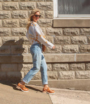 Woman walking past a brick wall on a sunny day, wearing a white patterned Dagny blouse with a paisley-shaped perforated design, jeans, and brown Bed Stu shoes.