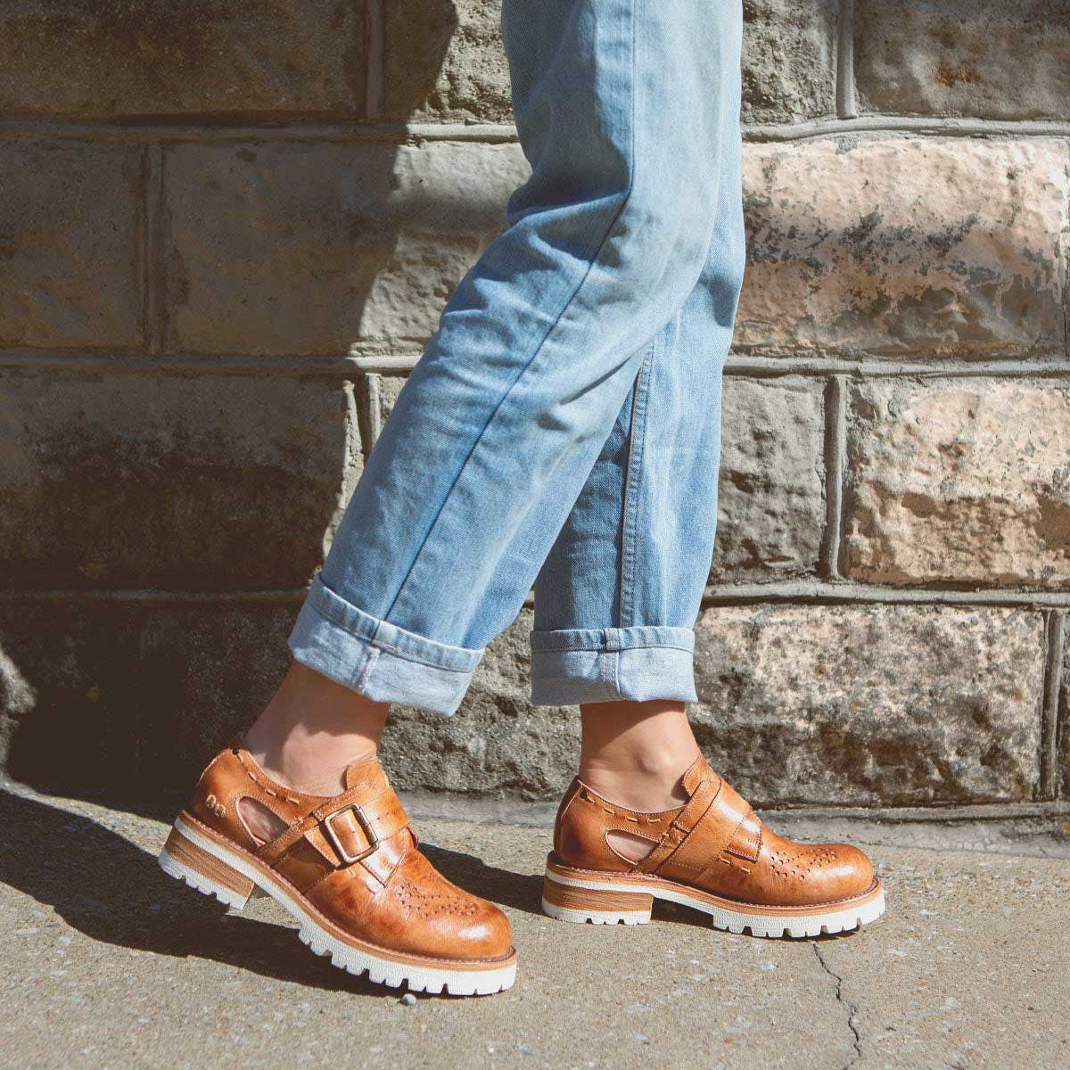 A person standing against a brick wall wearing brown Dagny brogues with a paisley-shaped perforated design and blue jeans by Bed Stu.