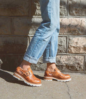 A person standing against a brick wall wearing brown Dagny brogues with a paisley-shaped perforated design and blue jeans by Bed Stu.
